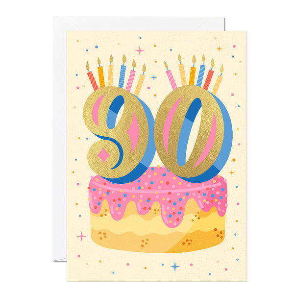 A 90th birthday card featuring a colourful illustration of a cake with the number 90 in gold foil, set against a beige background with stars and a 'Happy Birthday' message.