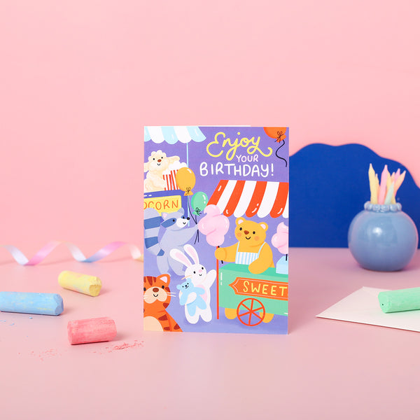 A colourful birthday card featuring cartoon animals at a carnival with balloons and a banner saying 'Enjoy your Birthday!'