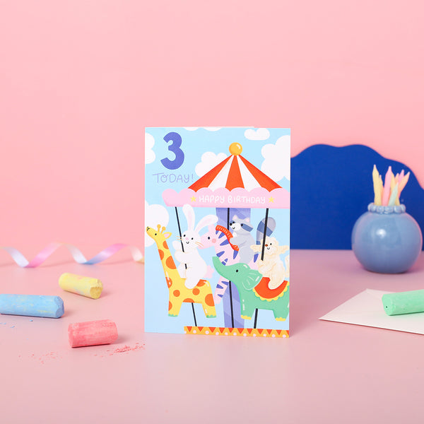 A colourful birthday card featuring an illustration of a carousel with a giraffe, rabbit, pig, and cat riding on it. The card indicates it's a 3rd birthday card and says 'Happy Birthday'.