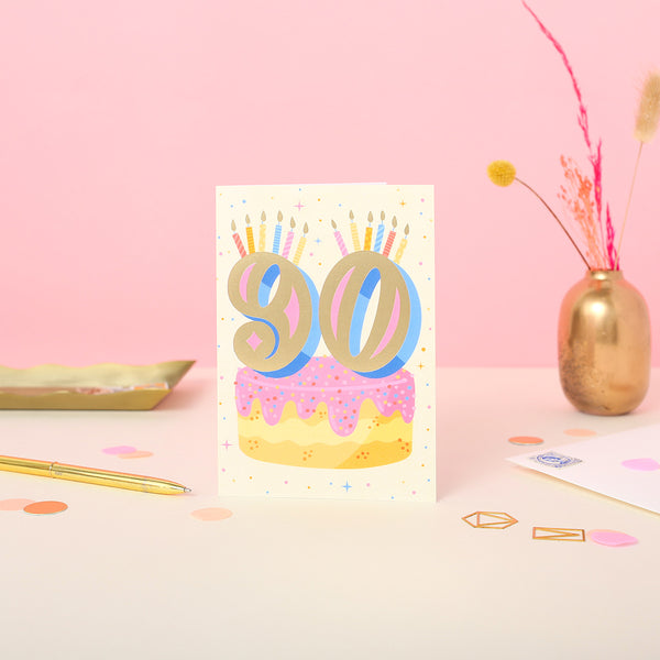A 90th birthday card featuring a colourful illustration of a cake with the number 90 in gold foil, set against a beige background with stars and a 'Happy Birthday' message.