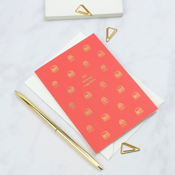 A greeting card with lots of illustrations of burgers and fries with the greeting 'we belong together' printed on a red card with gold foil