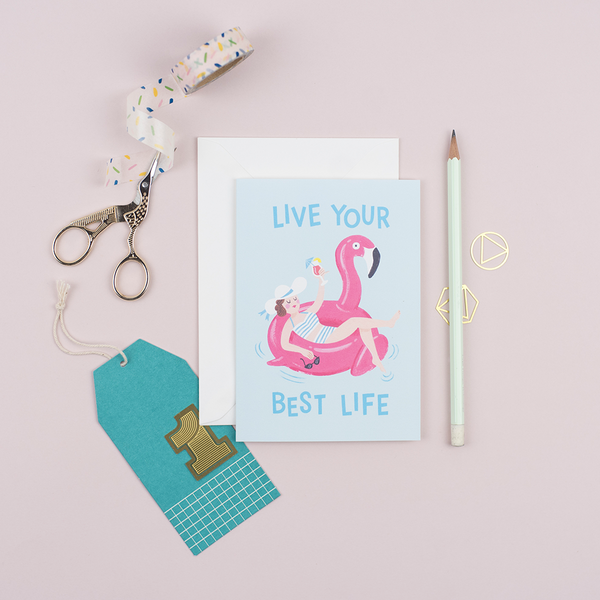 A birthday card that features an illustration of a woman holding a cocktail lounging on a flamingo pool float with a greeting that reads 'Live Your Best Life'