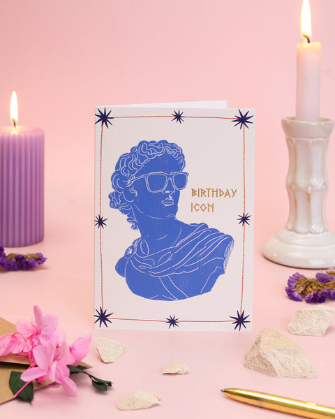 A chic cream birthday card displaying a silhouette of a blue Greek bust wearing sleek sunglasses, surrounded by delicate gold stars and elegant typography.