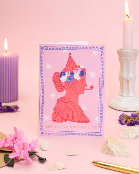 A playful terracotta-toned greeting card showcasing a Greek figure wearing a floral wreath and a striped party hat while blowing a celebratory party horn.