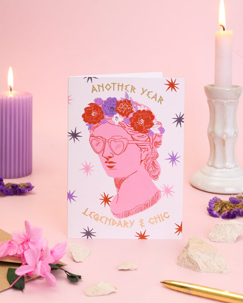 A fashion-forward birthday card featuring a pink classical bust wearing heart-shaped sunglasses and a rich crown of autumn-toned peonies and dahlias.
