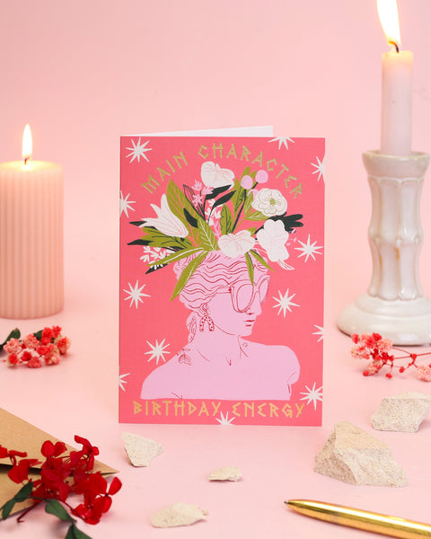 A maximalist coral birthday card illustrating a pink marble silhouette with drop earrings, sunglasses, and a spectacular crown of blooming lilies and wildflowers.