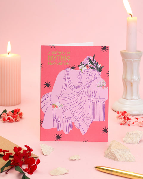 An artistic pink and coral birthday card featuring a draped Greek figure wearing a flower crown and leaning elegantly against a classical marble column.