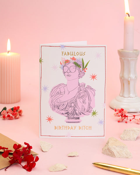 A sassy greeting card showing a pastel pink Greek bust accessorised with oversized black sunglasses and a large flower crown within a double border.