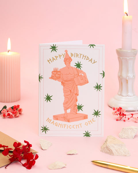 A modern birthday card featuring an illustrated classical Greek statue wearing a party hat and sunglasses while holding a towering birthday cake and a present.