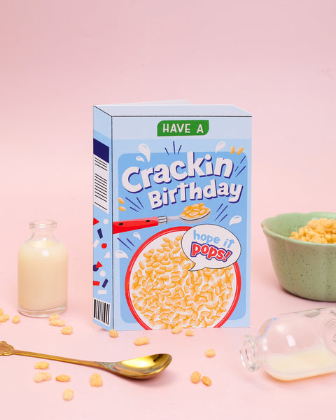 A die-cut greeting card featuring a puffed rice cereal box design on a bright blue background with a breakfast bowl and a 'hope it pops!' speech bubble.