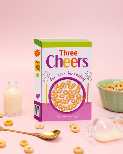 A die-cut birthday card shaped like a retro cereal box with an illustration of a breakfast bowl filled with hoops, celebratory sparklers, and bold typography.
