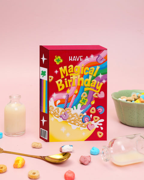 A die-cut birthday card depicting a whimsical marshmallow cereal box with a vibrant rainbow, magical charms, and colourful birthday candles.