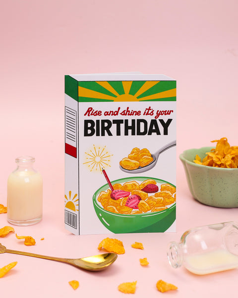 A die-cut birthday card illustrating a vibrant sunburst-themed cereal box with a crisp bowl of cornflakes, fresh strawberries, and a burning candle.