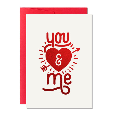 This is a Valentine's Day card that has a hand-lettered greeting that reads 'You and Me' with a cupid-inspired heart and arrow, printed with a sparkly red foil in the UK, paired with a red envelope.