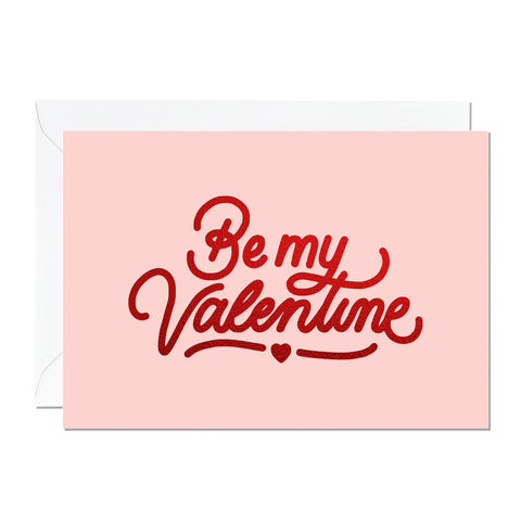 This is a Valentine’s Day card that says 'Be My Valentine'. It's printed with a light pink background and features hand lettering printed with a luxury sparkly red foil